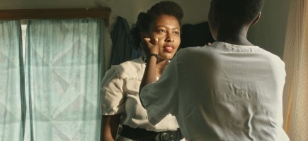 A woman in a white dress looks into the eyes of another woman who is touching her cheek.